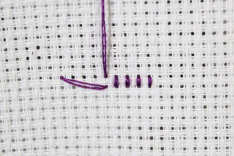 short row of cross stitch embroidery in purple thread on white aida cloth ending with an away knot view from the back