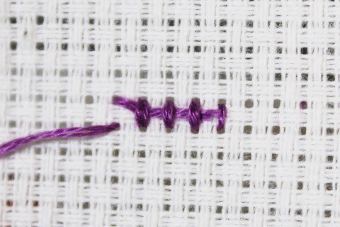 short row of cross stitch shown from the back with the tail end of the thread tucked under the stitches done in purple embroidery floss on white aida