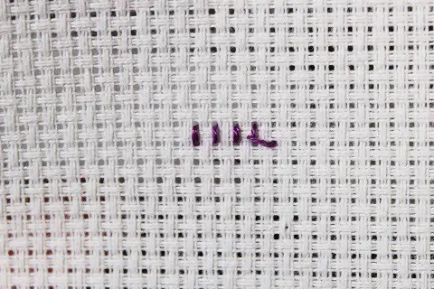 a short row of cross stitches viewed from the back showing the loop start with purple embroidery floss on white aida