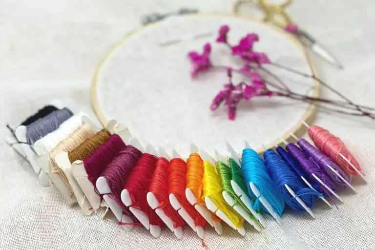 A rainbow of embroidery floss is splayed out under an embroidery hoop with linen fabric and a sprig of delicate, pink flowers with a pair of gold embroidery scissors next to the hoop