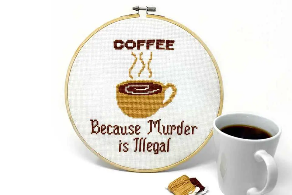 Bamboo embroidery hoop containing cross stitch project that reads Coffee, Because Murder is Illegal, with a beige coffee mug, all on a white background